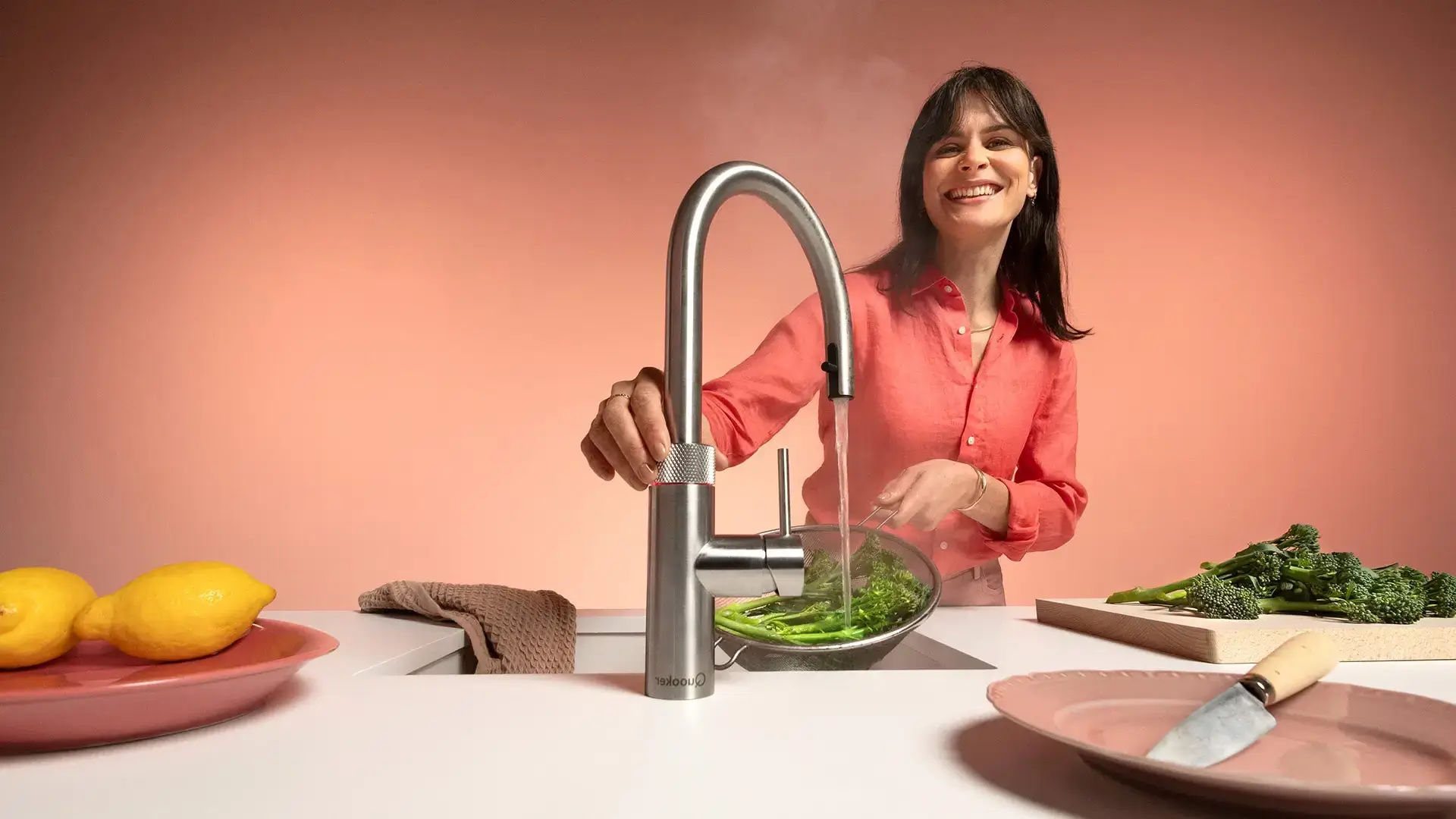 Frau wäscht grünes Gemüse in einem Edelstahl-Spülbecken mit einem modernen Wasserhahn.